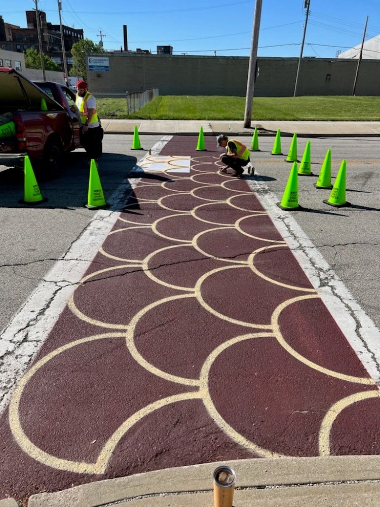 Volunteers install decorative crosswalk on Payne Avenue. A painter is protected by green cones and a construction truck while painting in the middle of the crosswalk.