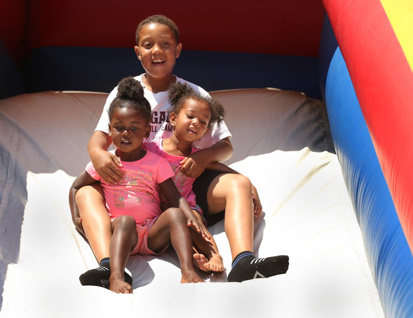 Three young children sitting together at the top of an inflatable slide, smiling and getting ready to slide down on a sunny day.