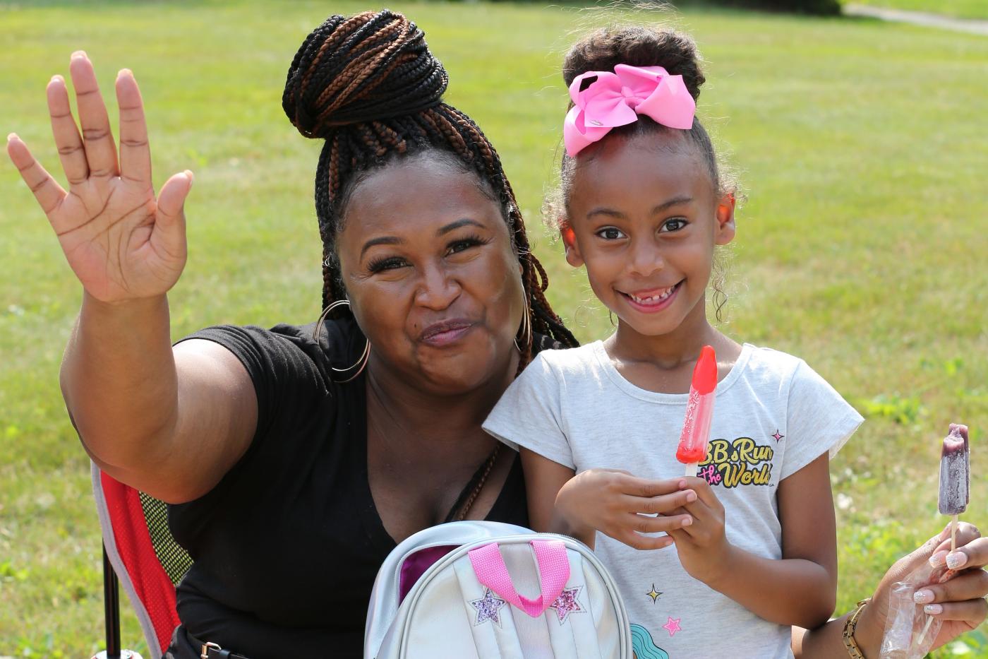 Woman and young girl smiling outdoors on a sunny day, waving at the camera and holding colorful popsicles while sitting in a grassy park setting.