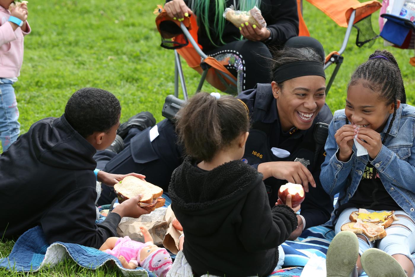 Police officer smiling and sitting on the grass with a group of children during an outdoor picnic, as the kids eat sandwiches and fruit and interact together in a park setting.
