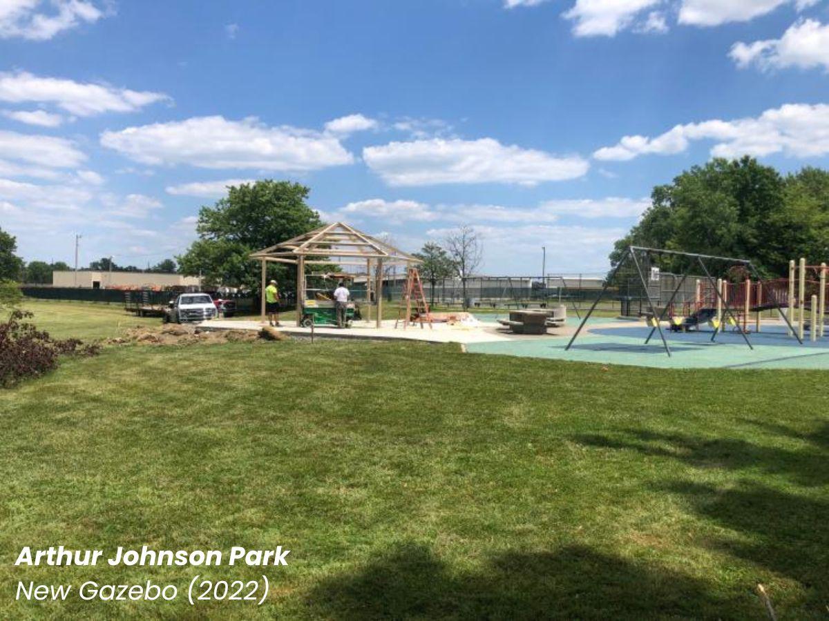 Arthur Johnson Park new gazebo under construction in the middle of a grassy lawn with a few large trees and nearby playground.