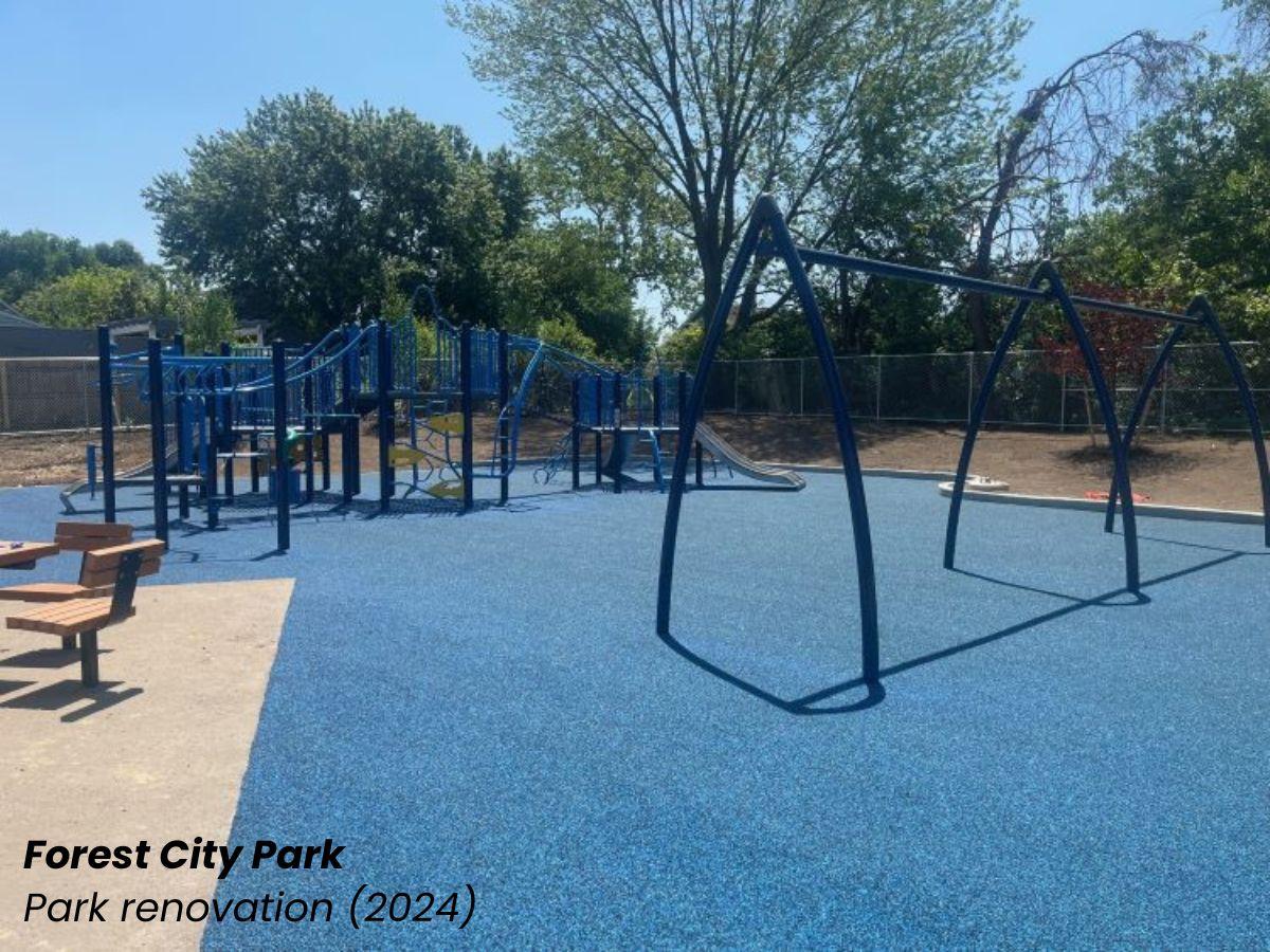 A renovated playground at Forest City Park features blue rubber safety surfacing, modern play equipment, swings, and benches surrounded by trees on a sunny day.