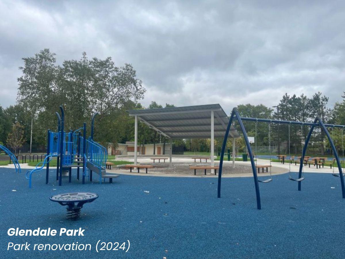 A renovated playground at Glendale Park includes blue rubber safety surfacing, swings, climbing structures, shaded seating areas, and open recreation space under a cloudy sky.