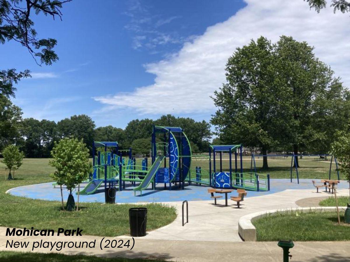 A modern blue and green playground with slides, swings, benches, and accessible pathways sits in a grassy park under a partly cloudy sky.