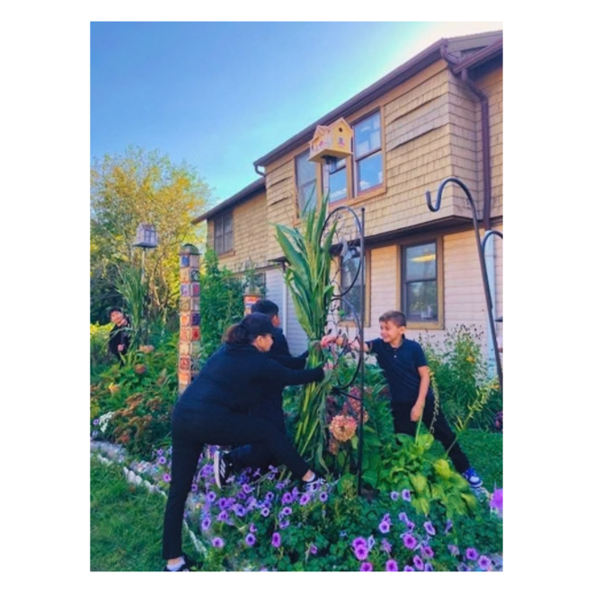 A Social Support Services staff member and a young person work together in a colorful garden, arranging tall plants near a birdhouse and decorative garden features beside a community building.