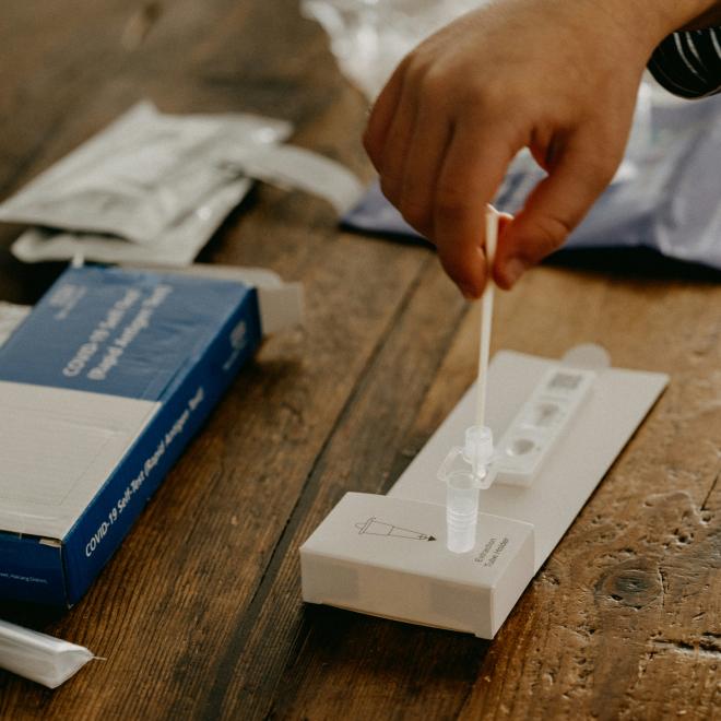A hand holding a swab into a COVID-19 at home testing vial. Other testing material is also on the table.